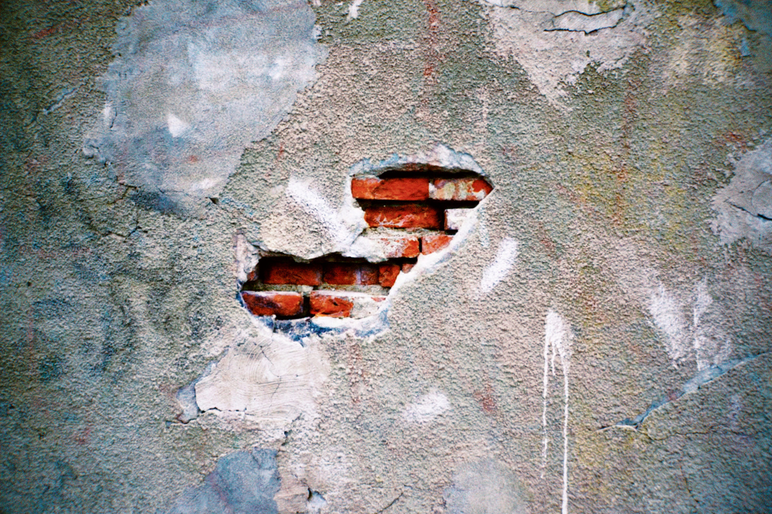 Photo of a concrete wall with a hole in it revealing a red brick wall beneath