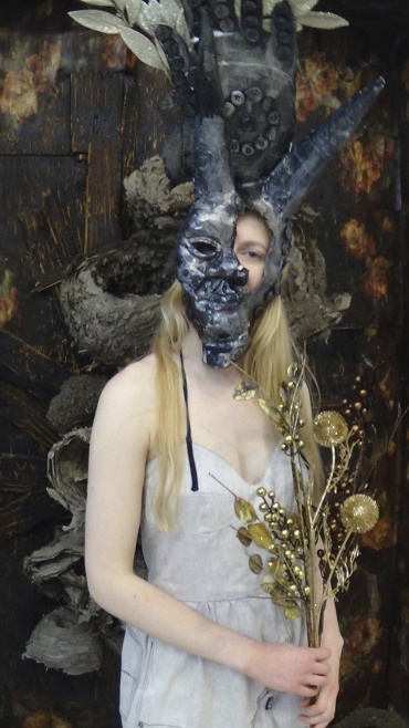 Photo of a woman in a white dress wearing a horned mask over half her face and holding a bouquet of gold metal flowers