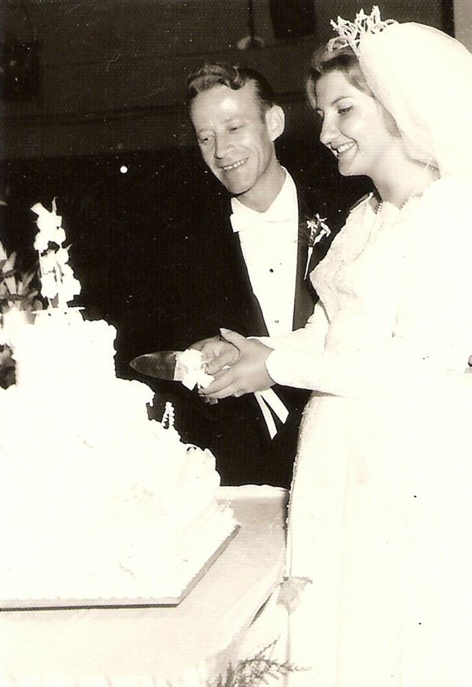 Old photo of a man and woman in wedding attire smiling and cutting a tiered cake