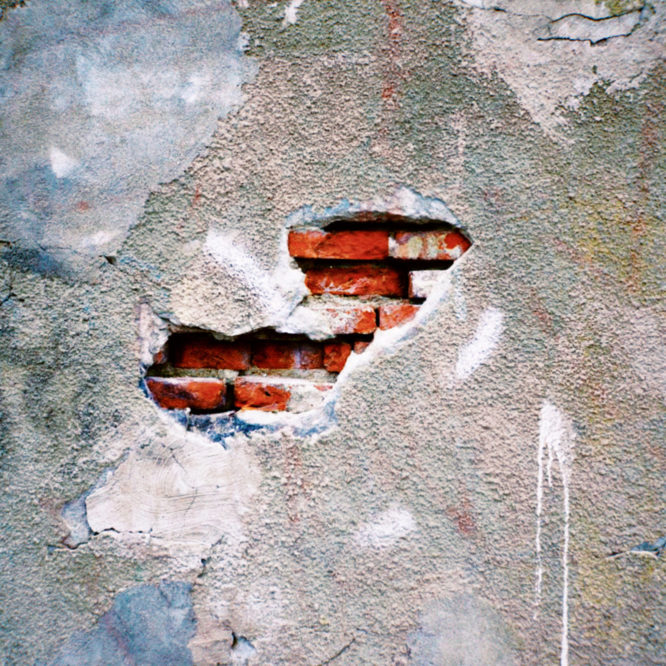 Photo of a concrete wall with a hole in it revealing a red brick wall beneath